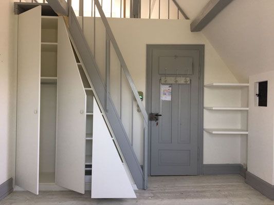 Weisser Einbauschrank unter der grauen Treppe. Graue Tür mit Regalen an der Wand.