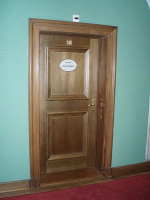 Holztür mit Goldrand, beschriftet mit „16“. Ein ovales Schild ist an der Tür angebracht. Hellgrüne Wände und roter Boden.