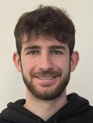 Mann mit dunklen Haaren und Bart, der einen schwarzen Kapuzenpullover trägt, lächelt in die Kamera.