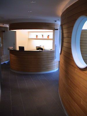 Gebogener Empfangstresen aus Holz in einem modernen Interieur mit rundem Fenster und dunklem Boden.