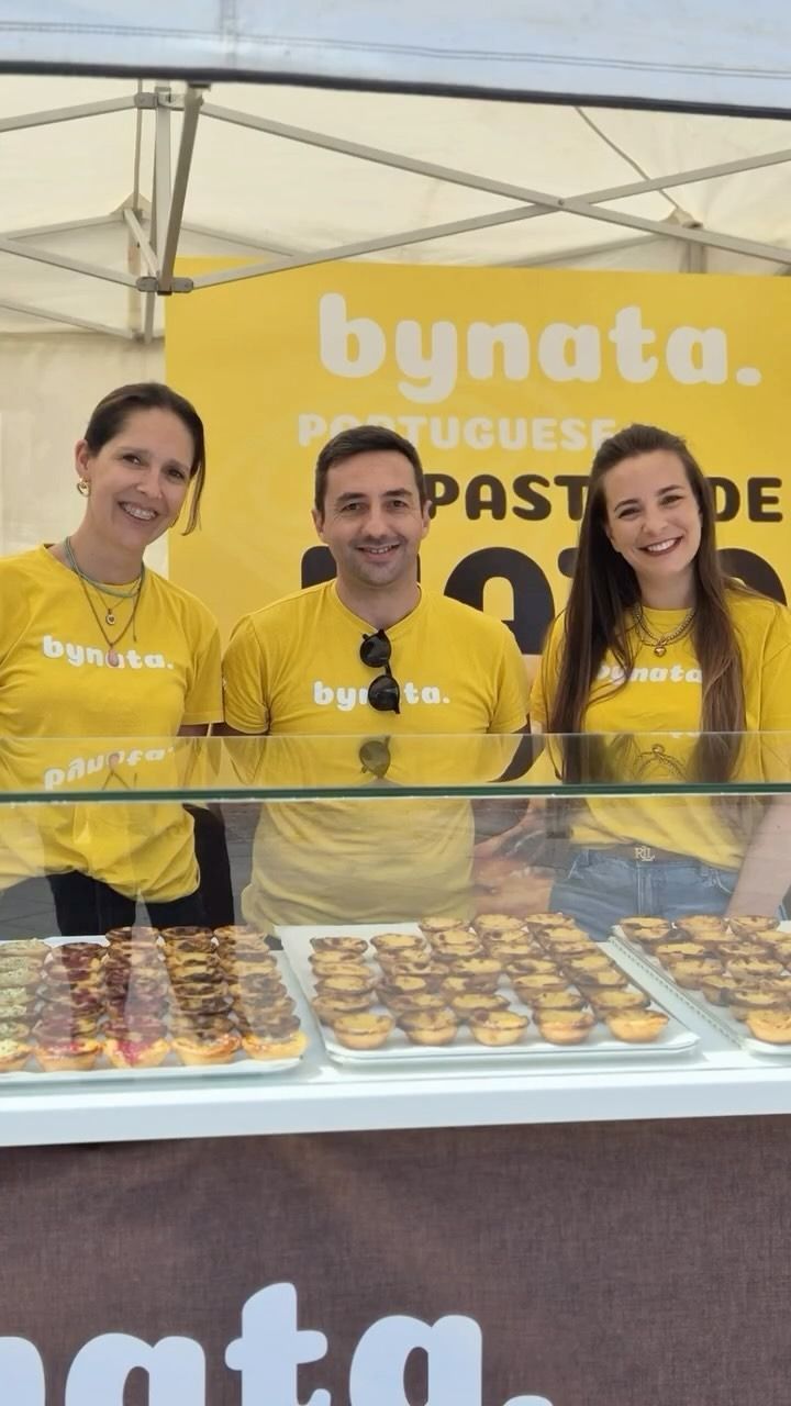 Un grupo de personas de pie detrás de una exhibición de pasteles frente a un cartel que dice bynata