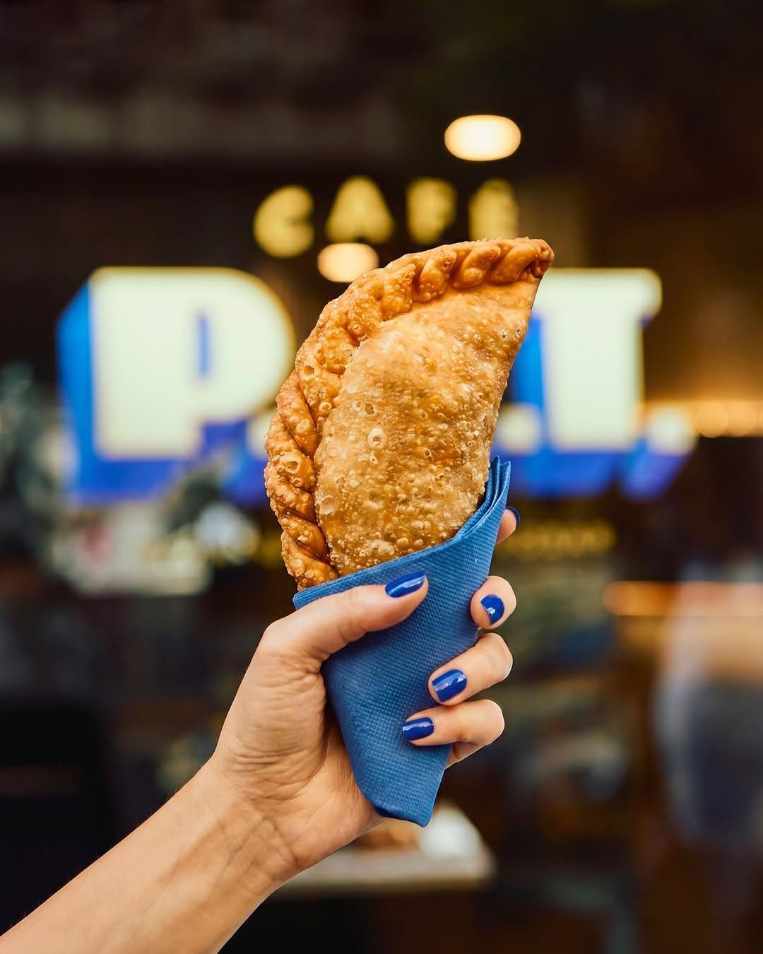 Una mano sostiene un pastelito frito dorado envuelto en una manga azul, con un letrero de cafetería borroso al fondo.