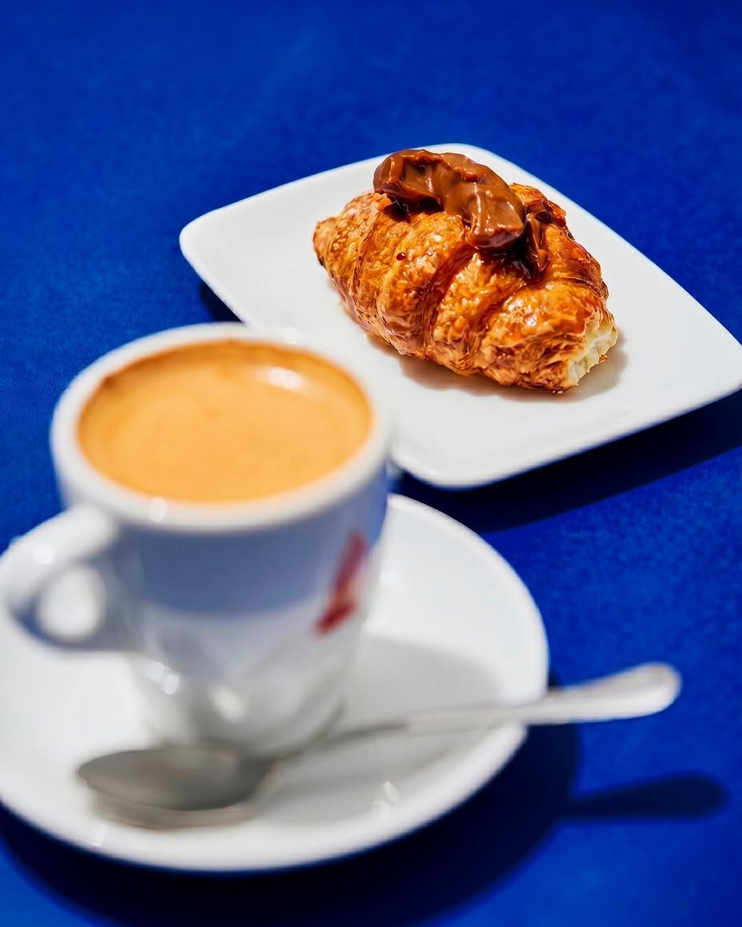 Taza de café y cruasán sobre platos blancos, frente a un mantel azul.
