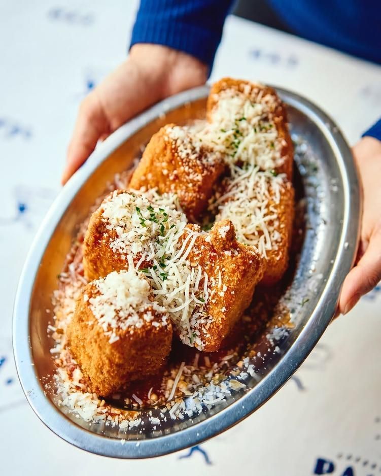 Manos sosteniendo una bandeja de aperitivos empanizados con salsa y cubiertos con queso rallado.