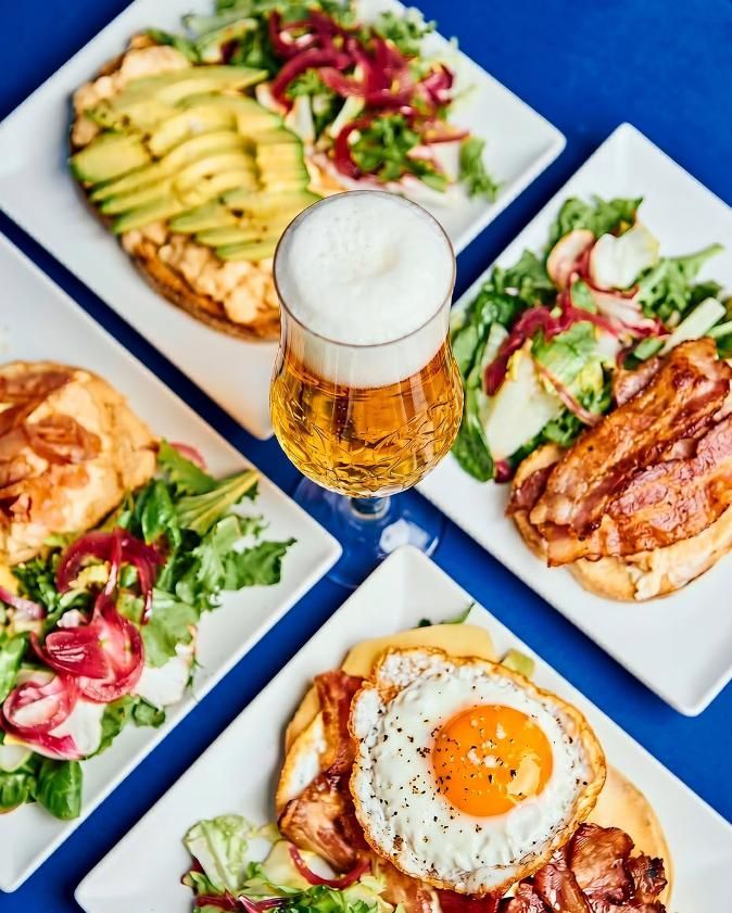 Vista aérea de un brunch con sándwiches, ensalada, tostadas de aguacate y un vaso de cerveza servidos en un mantel azul.