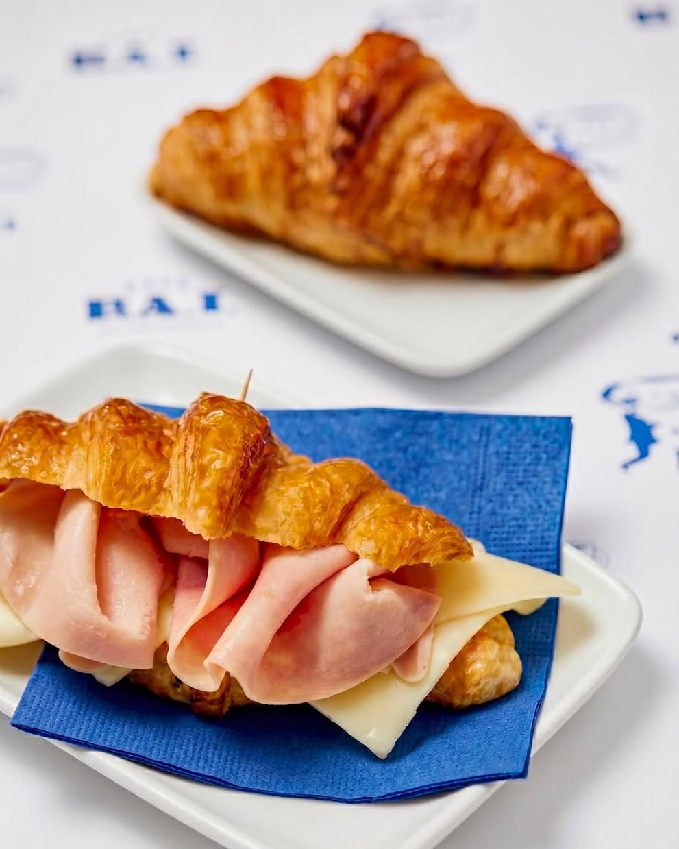 Dos sándwiches de croissant en platos blancos, uno de ellos con jamón y queso sobre una servilleta azul.