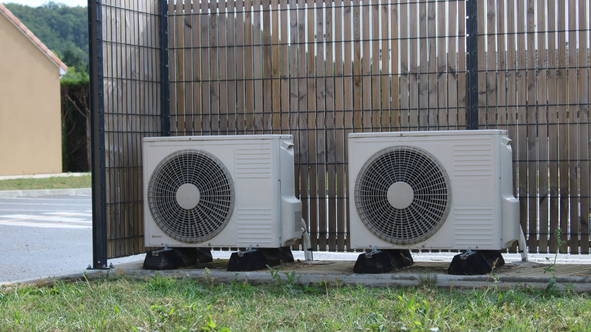 Deux unités de climatisation extérieures beiges, placées côte à côte sur des dalles de béton, devant une clôture verticale en bois assurant l'intimité.