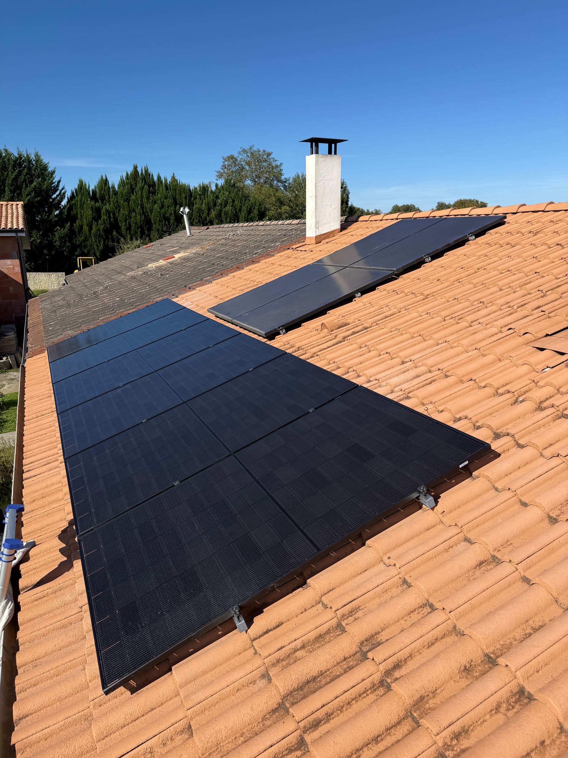 Des panneaux solaires noirs sont installés sur un toit de tuiles en terre cuite, à côté d'une cheminée blanche, sur fond de ciel bleu clair.