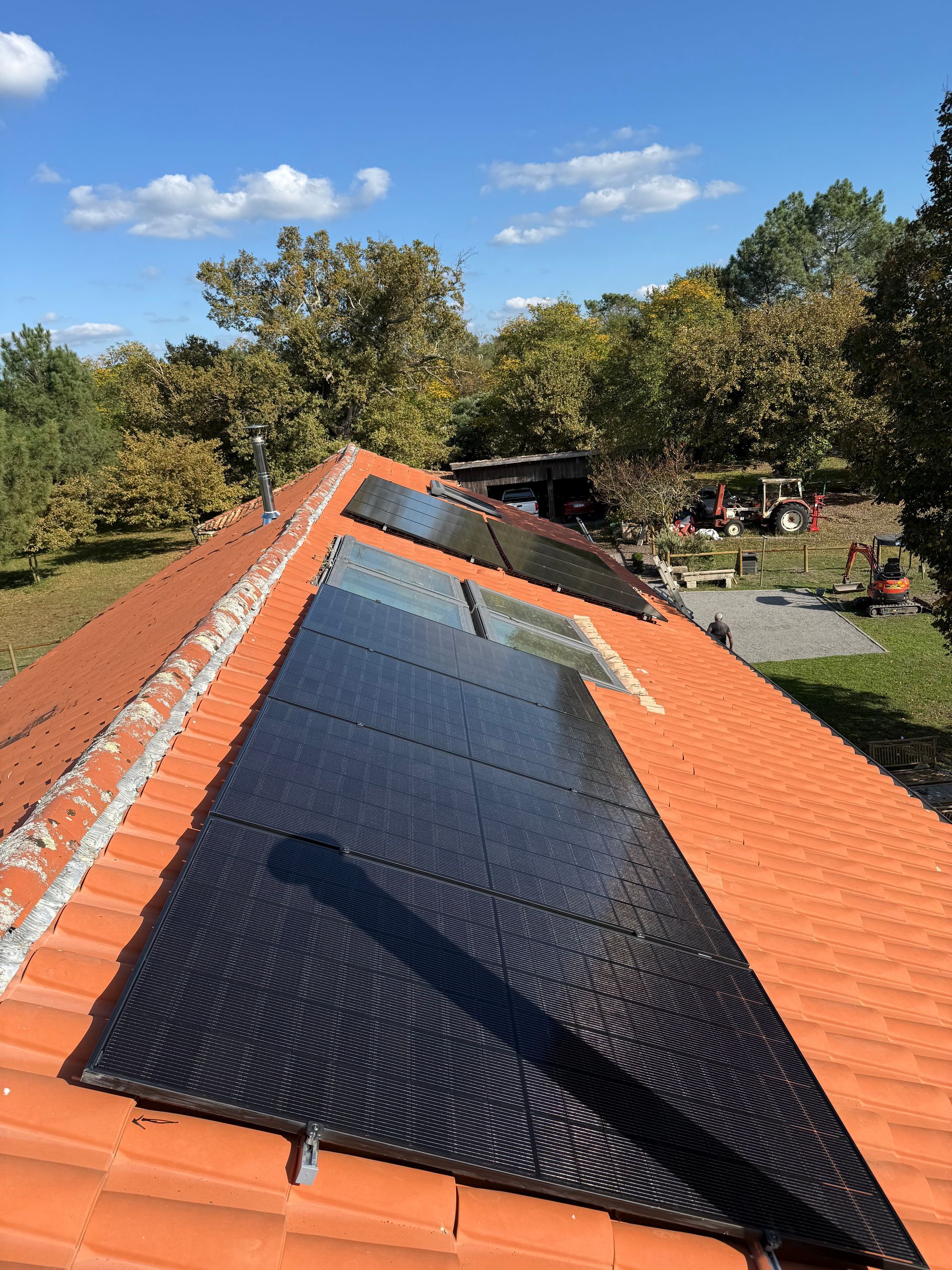 Une rangée de panneaux solaires noirs installés sur un toit de tuiles rouges, avec des arbres et un ciel bleu ensoleillé en arrière-plan.