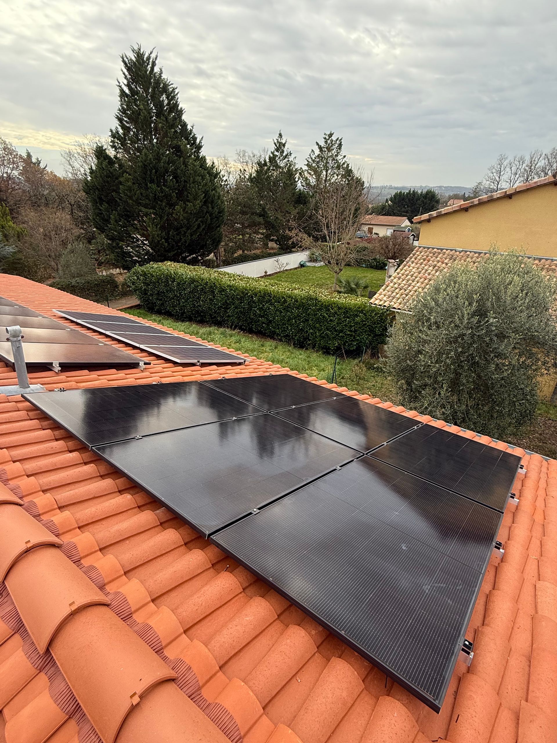 Panneaux solaires installés sur un toit de tuiles en terre cuite, avec un jardin et des arbres en arrière-plan.