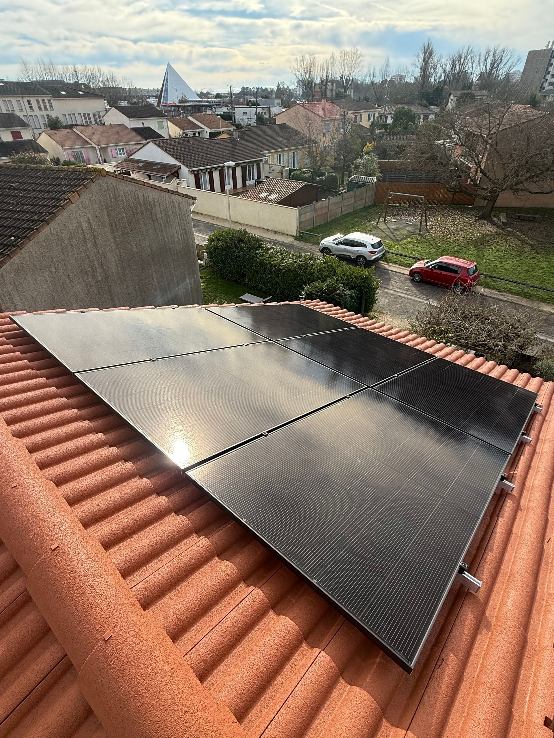 Un ensemble de panneaux solaires installé sur un toit en pente recouvert de tuiles en terre cuite, donnant sur une rue de banlieue bordée de maisons et de voitures.