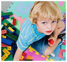 Niño sentado sobre una colorida alfombra de espuma con forma de rompecabezas y bloques LEGO, mirando hacia arriba mientras sostiene un bloque rojo.