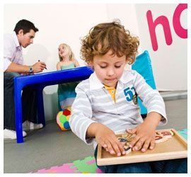 Un niño juega con un rompecabezas de madera en un aula colorida, con adultos al fondo.