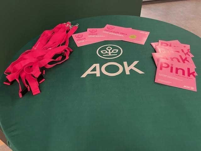 Pinkfarbene Schlüsselbänder, Karten und „Pink“-Karten auf einem grünen Tisch mit dem AOK-Logo.