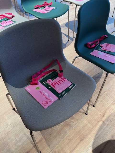 Graue und grüne Stühle mit Namensschildern und rosa Schlüsselbändern in einem Veranstaltungsort mit Holzboden.