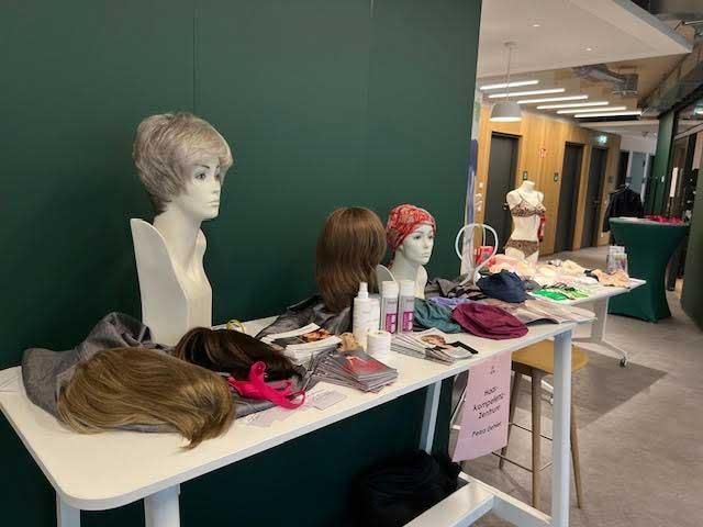 Perücken auf einem weißen Tisch vor einer grünen Wand. Schaufensterpuppenköpfe mit Perücken und verschiedenen Haarpflegeprodukten.