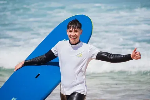 Un hombre sostiene una tabla de surf en la playa y hace un gesto de aprobación con el pulgar hacia arriba.