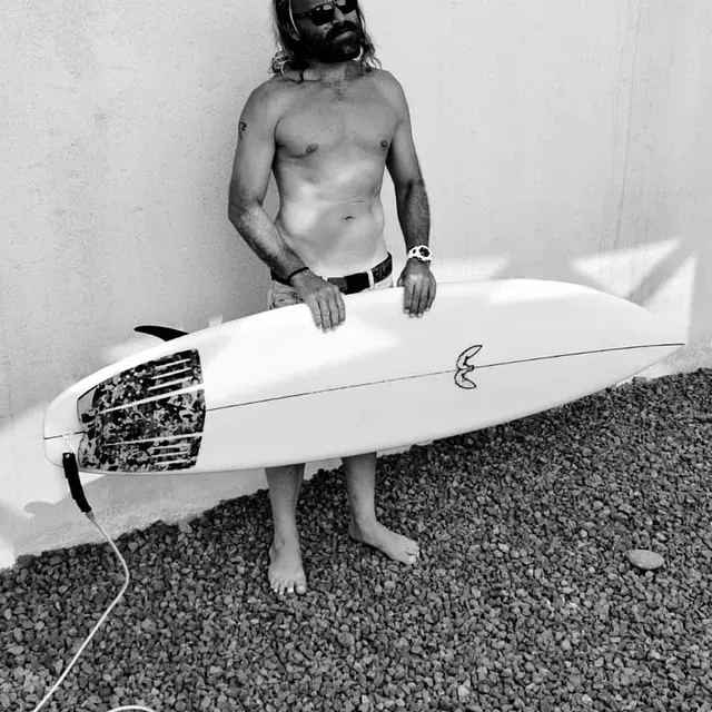 Un hombre sin camisa sostiene una tabla de surf con la letra e escrita en ella.