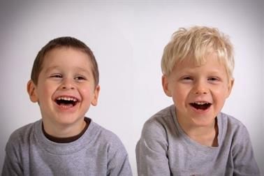 Zwei lachende Jungen, braunes und blondes Haar, graue Hemden, weißer Hintergrund.