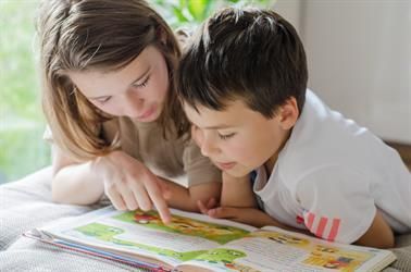 Mädchen zeigt auf ein Buch, während es mit einem Jungen liest, beide liegen konzentriert im Haus.