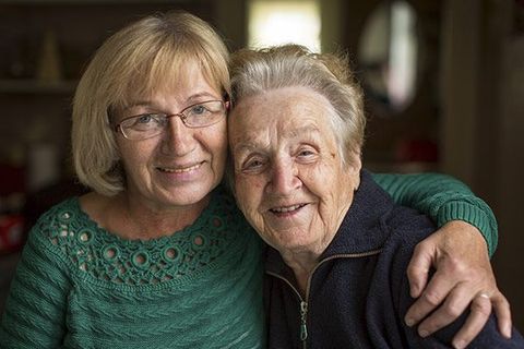 Frau umarmt eine ältere Frau, beide lächeln. Die jüngere Frau trägt eine Brille und einen grünen Pullover.