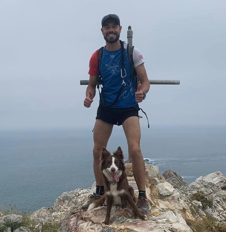 Hombre y perro en una cima rocosa, pulgares arriba. Océano al fondo, cielo nublado. El hombre viste ropa deportiva.