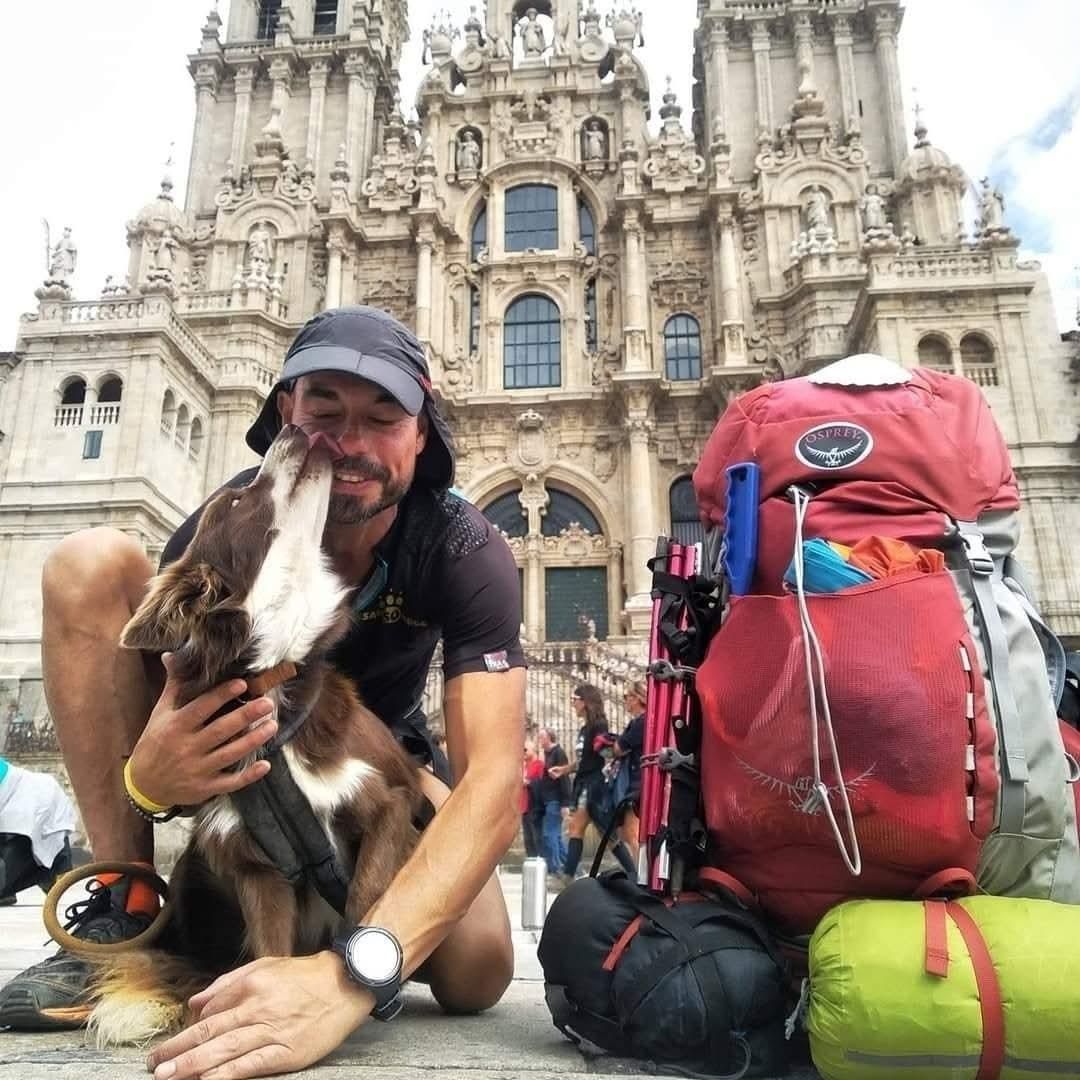 Hombre y perro frente a una catedral; hombre abraza a perro, mochila roja a su lado.