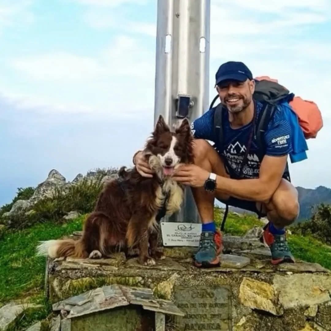Hombre y perro en la cima de una montaña con una cruz de metal. El hombre sonríe y el perro mira a la cámara.
