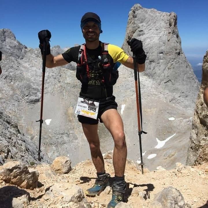 Hombre con bastones de senderismo en una montaña, vistiendo ropa para correr, extendiendo los brazos en señal de victoria.