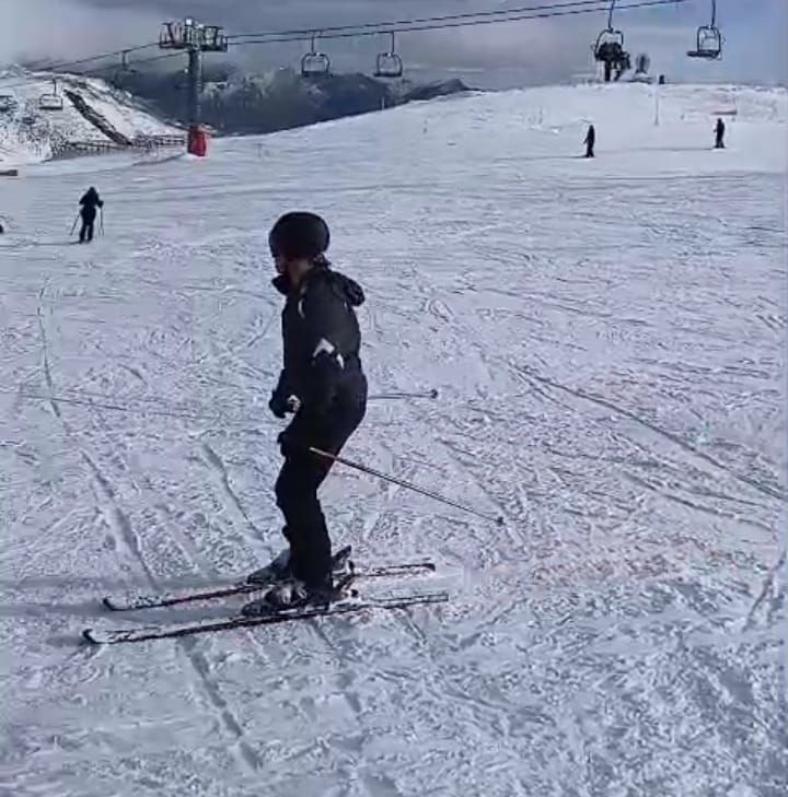 Esquiador con equipo negro en una pendiente cubierta de nieve; telesquí al fondo.