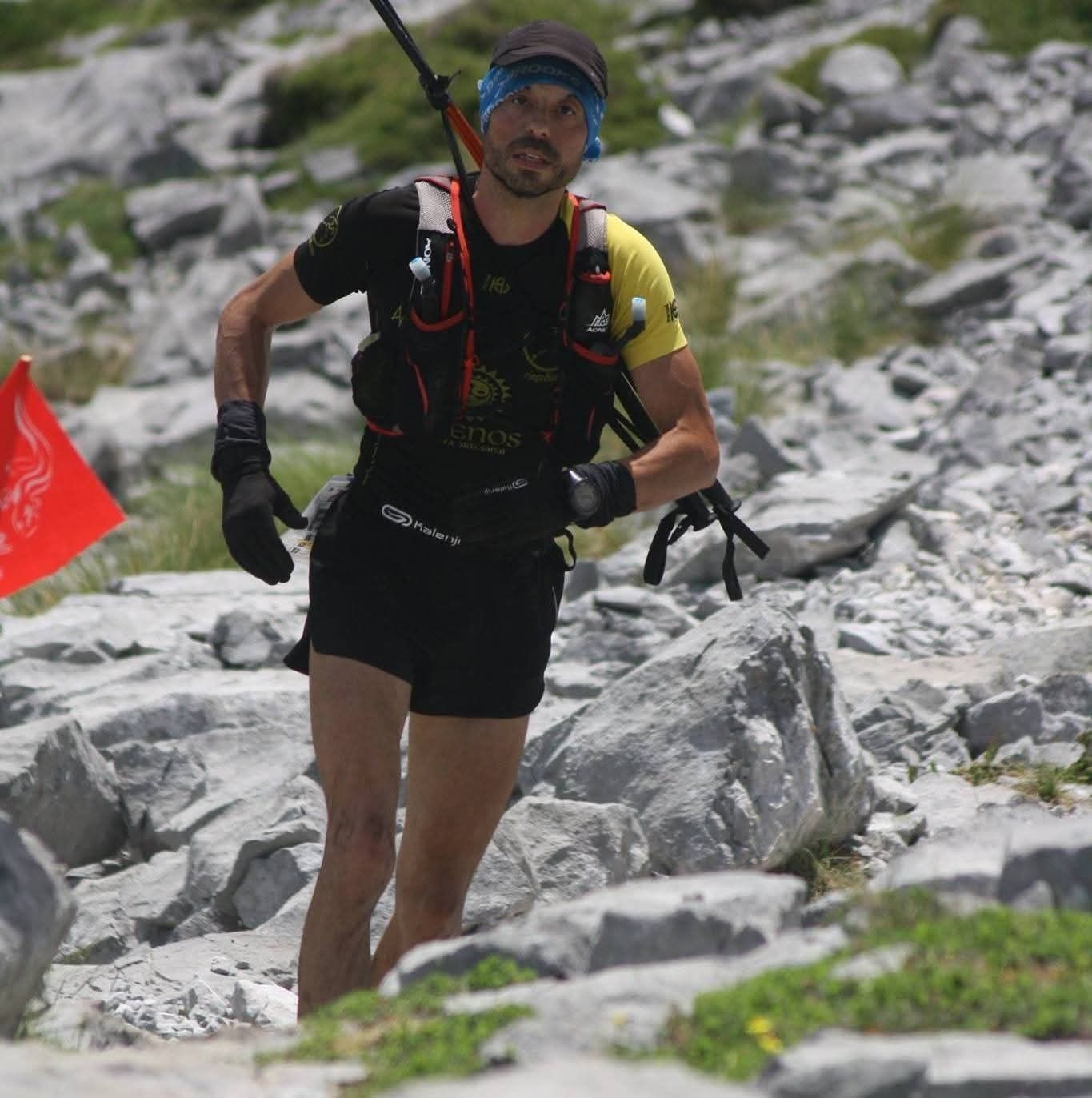Corredor con pantalones cortos y chaleco negros, usando bastones de trekking.
