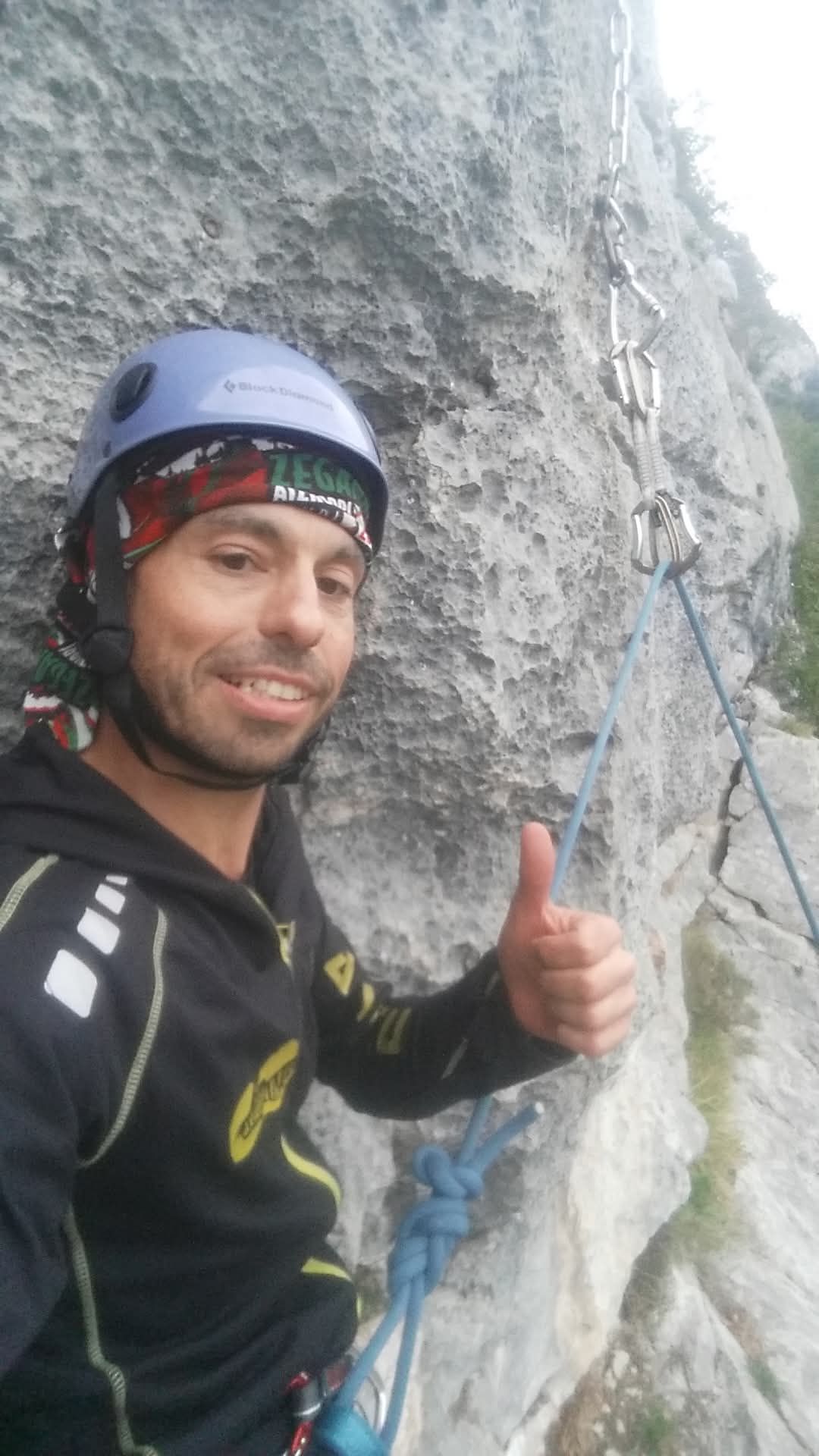 Un escalador con casco azul levanta el pulgar en una pared rocosa. Se ven una cuerda de seguridad y mosquetones.