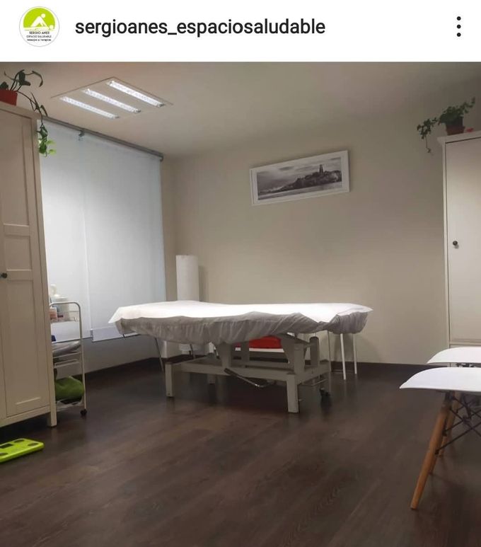 Una sala de terapia de masajes con una mesa de tratamiento, gabinetes blancos y una ventana con persianas.