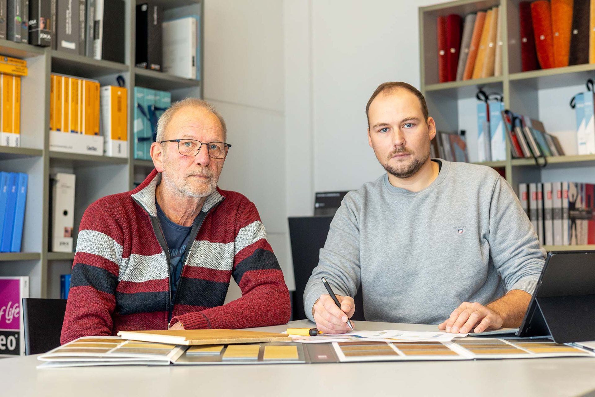 Zwei Männer sitzen an einem Tisch vor einem Bücherregal.