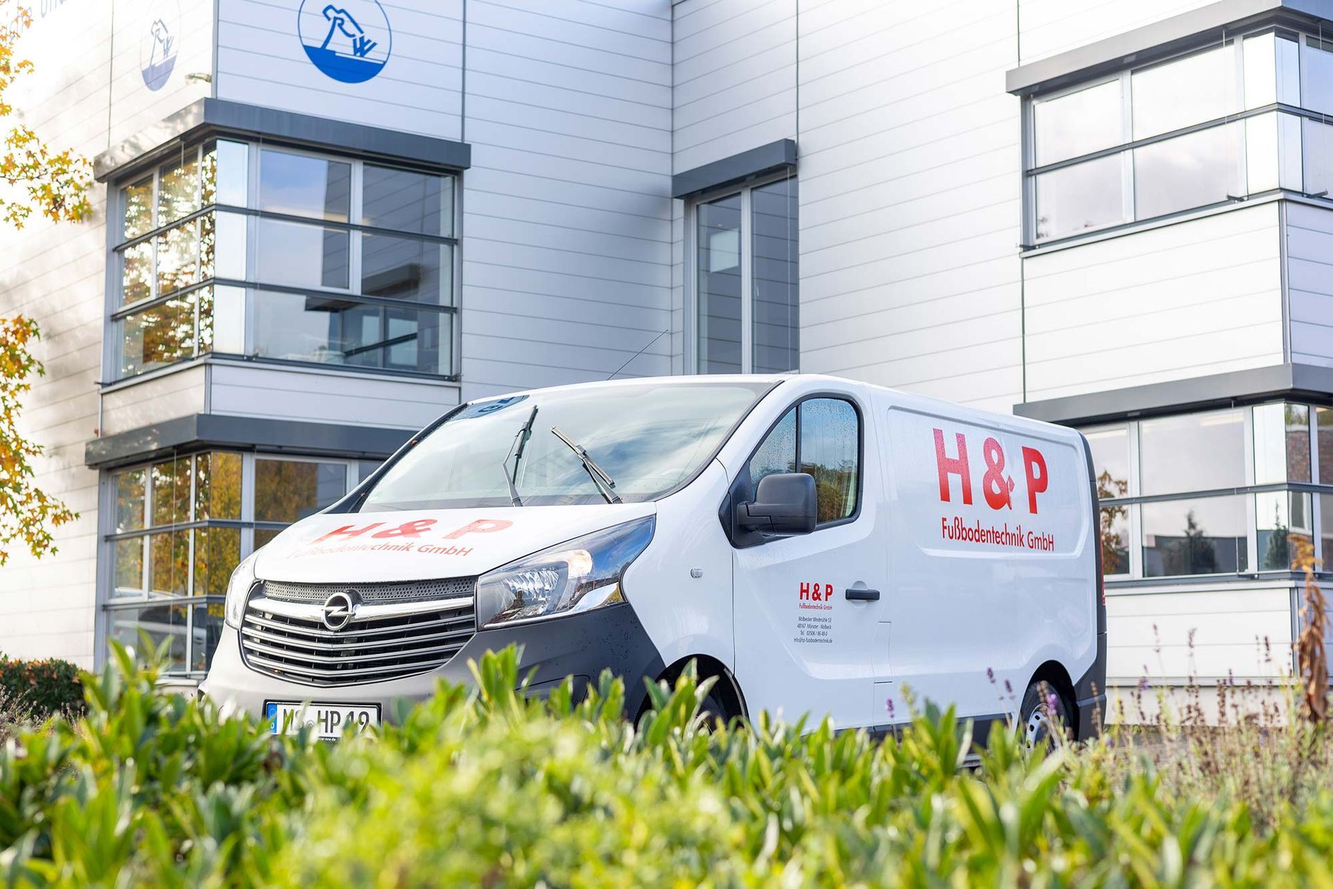 Ein weißer H&P-Lieferwagen parkt vor einem modernen Bürogebäude mit Grünflächen im Vordergrund.