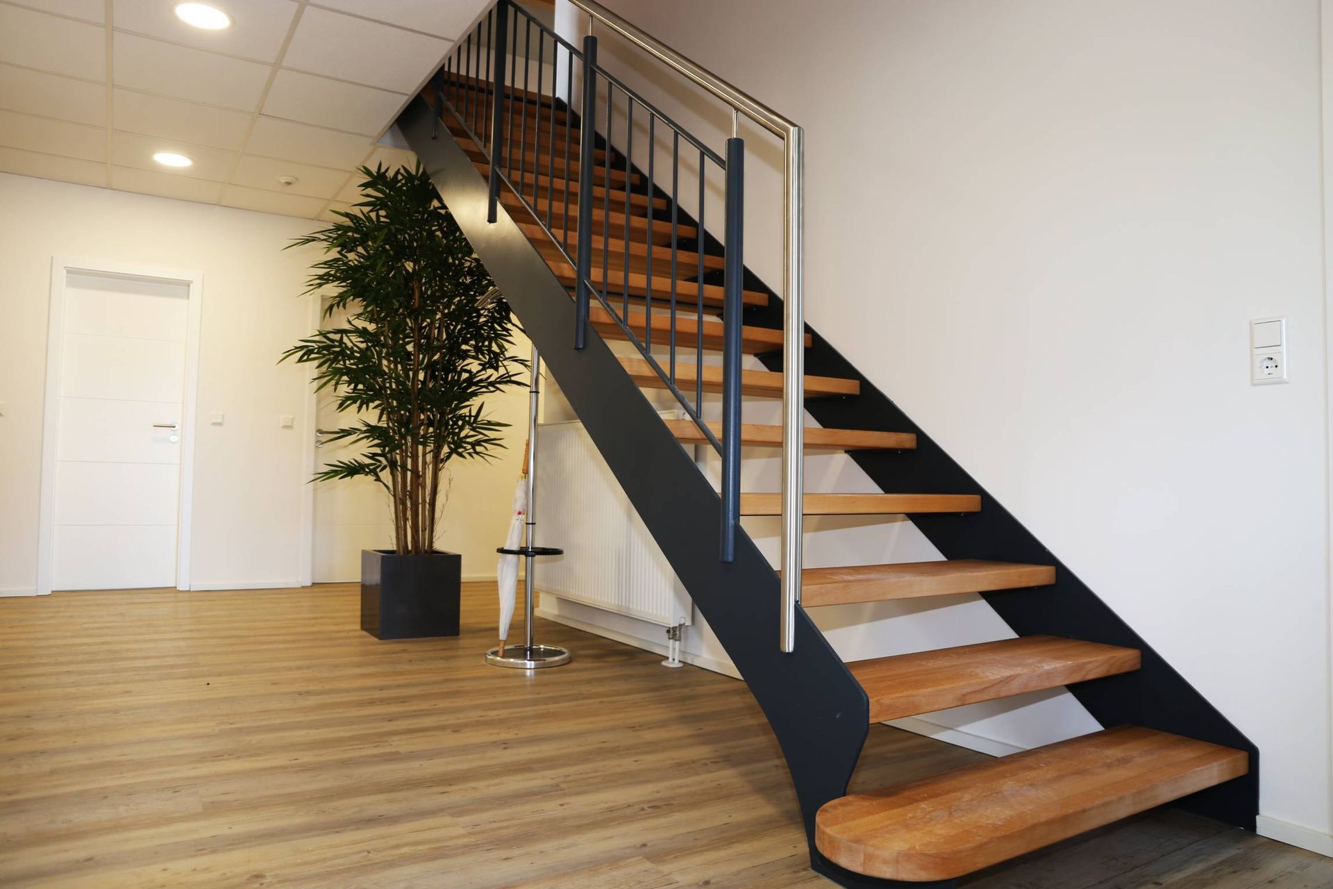 Escalier moderne à l’intérieur, avec une rampe noire, dans une pièce lumineuse dotée d’un parquet et d’un arbre en pot