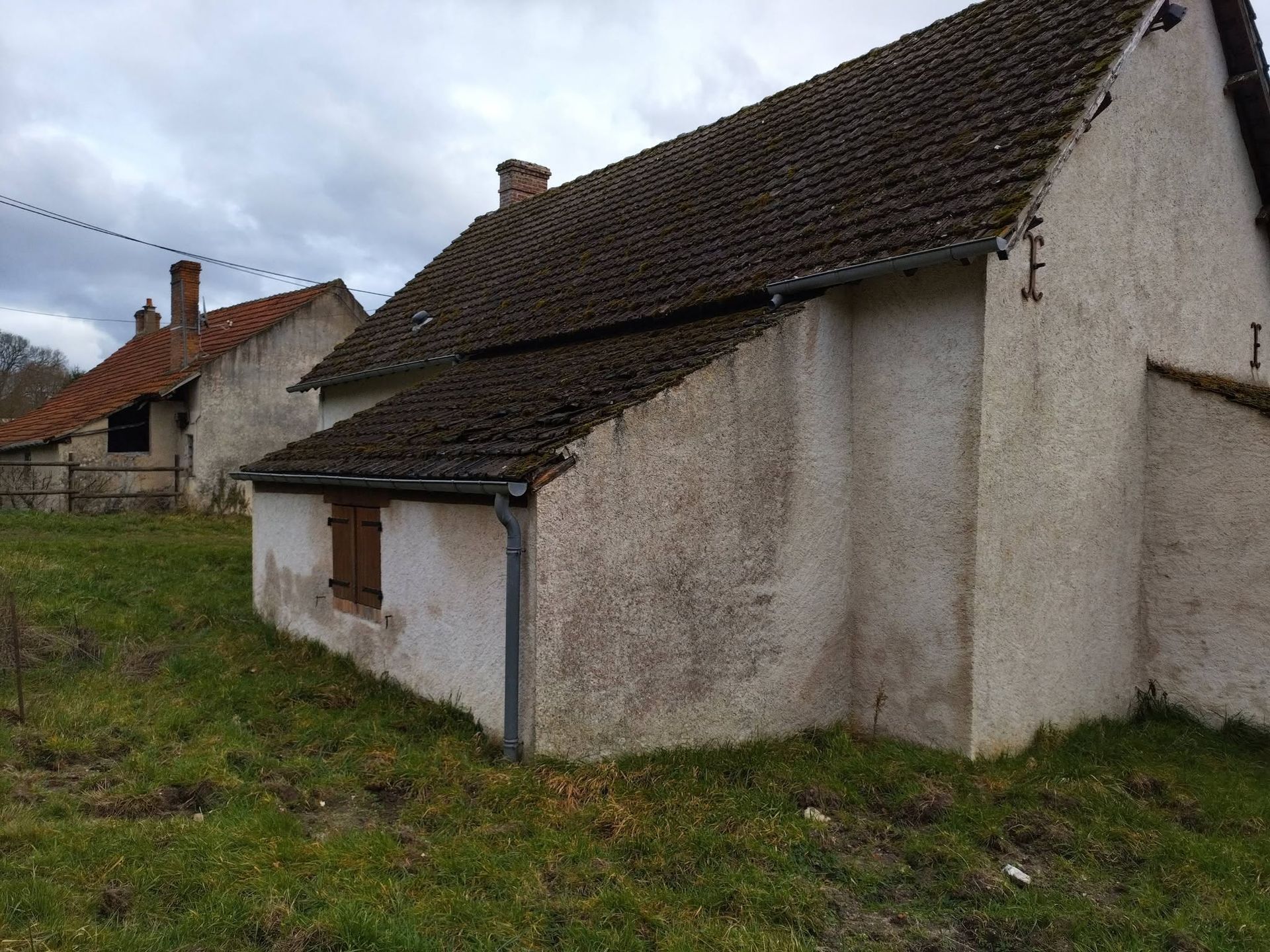 Vieille maison en pierre blanche au toit de tuiles foncées, dans une cour rurale herbeuse sous un ciel nuageux