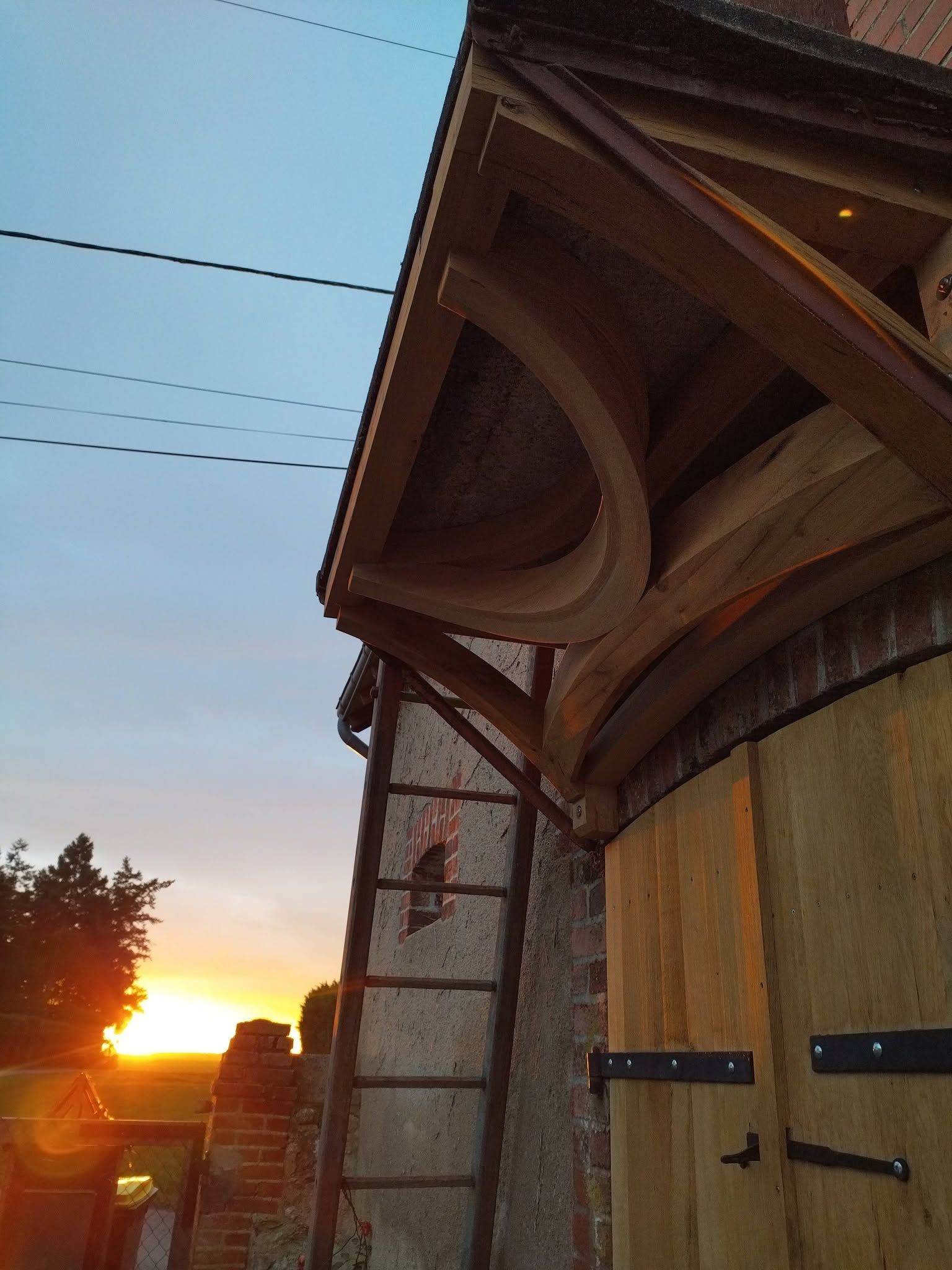 Coucher de soleil sur un bâtiment rustique en bois, avec un toit cintré et des lignes de service en silhouette