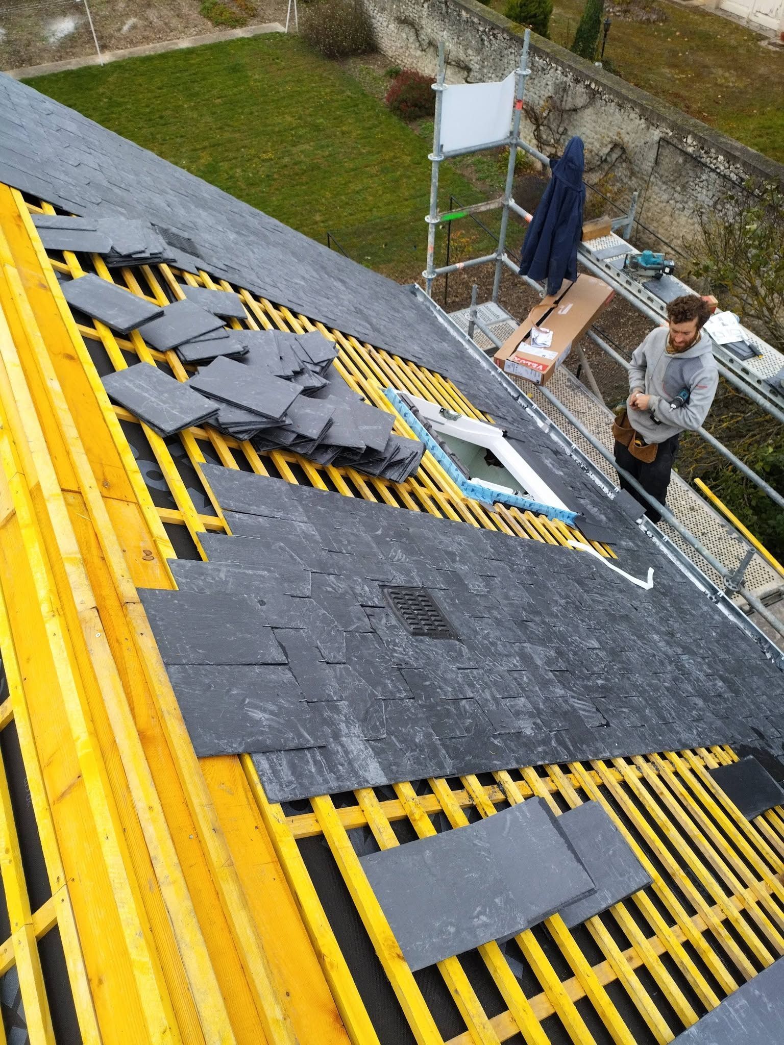 Un couvreur travaille sur un toit en pente couvert de bardeaux gris, avec des garde-corps de sécurité et un échafaudage jaune
