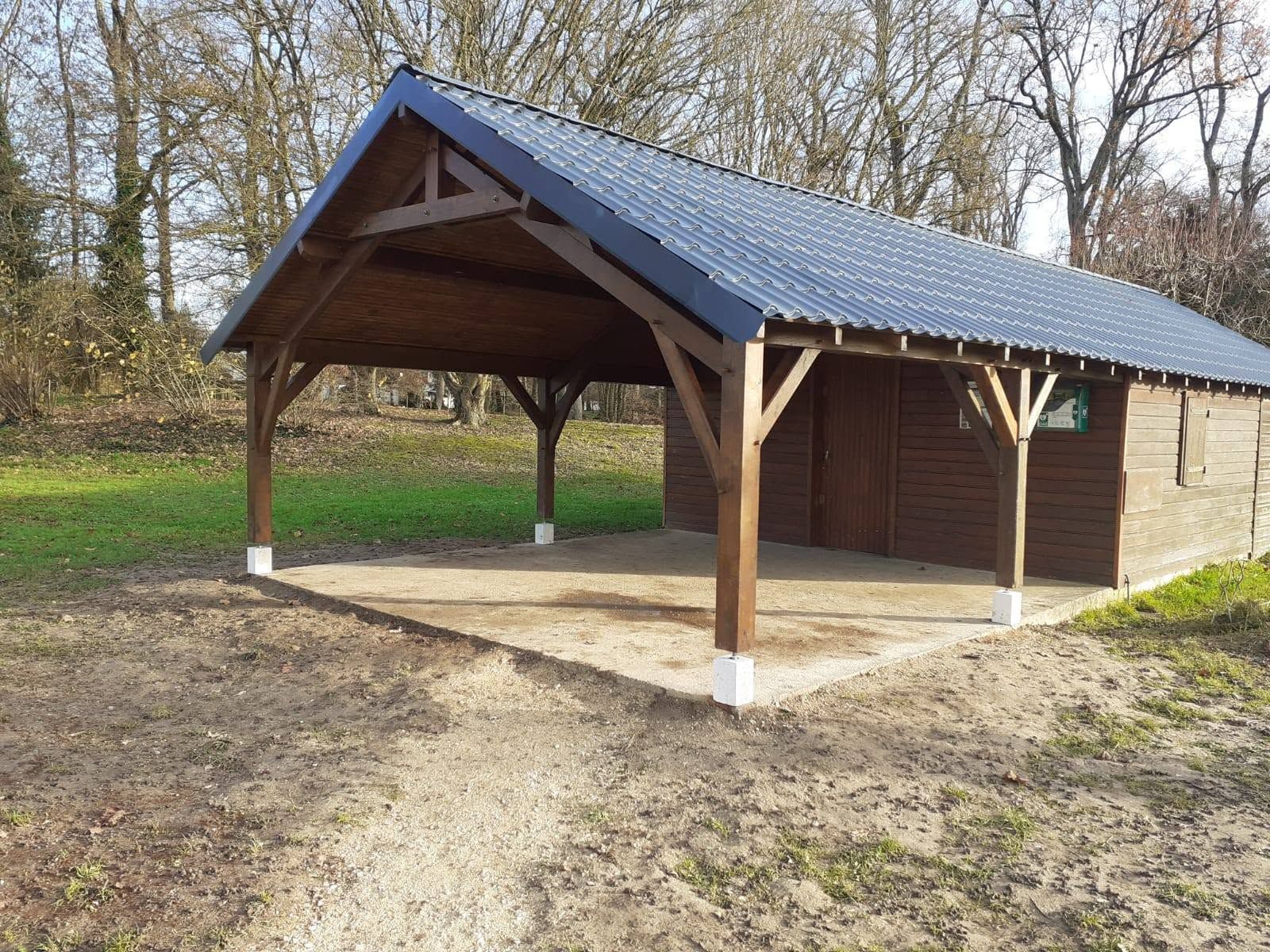 Pavillon en bois ouvert sur les côtés, avec un toit bleu et un chemin de terre dans un parc boisé