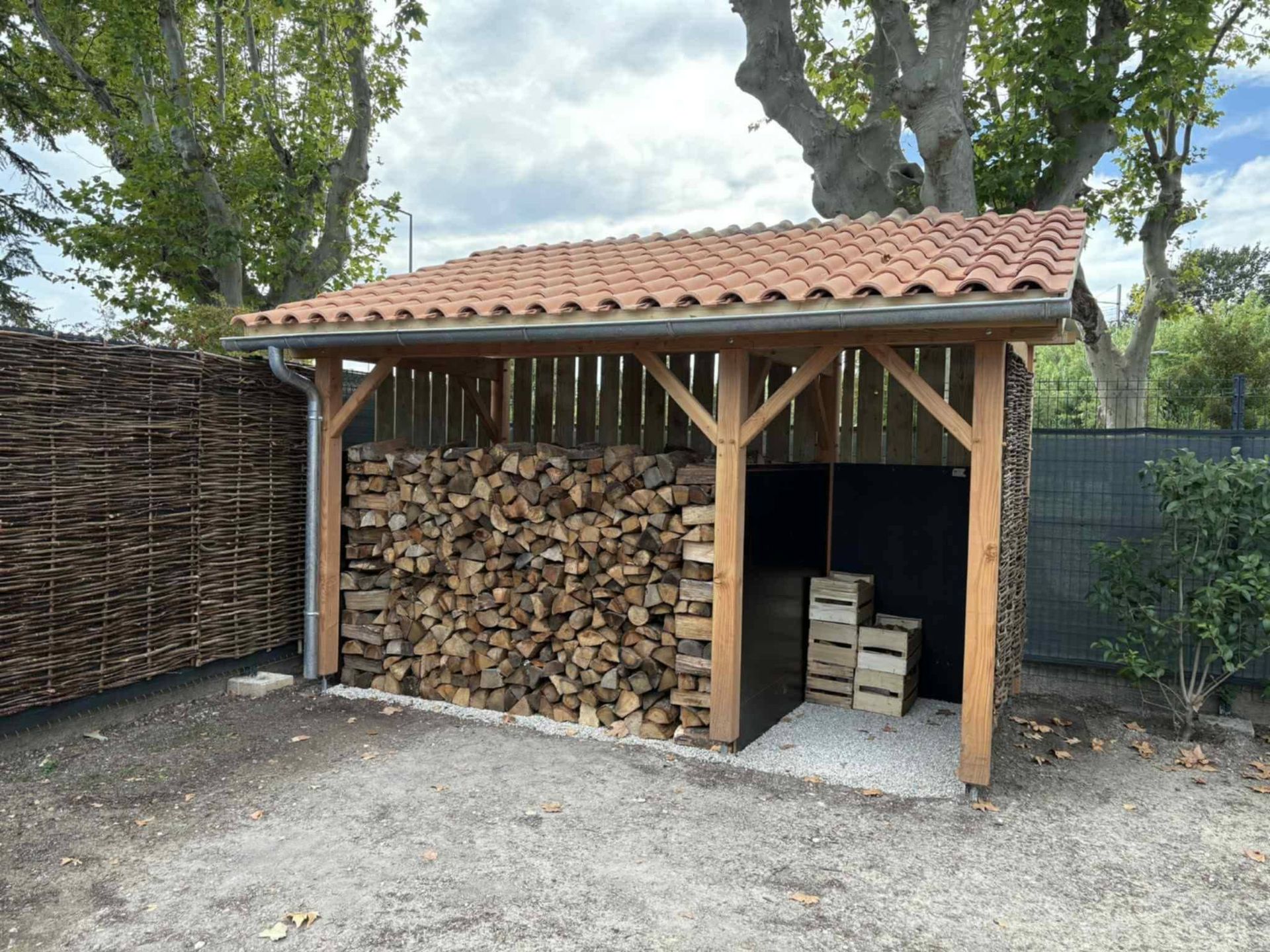 Abri à bois avec des bûches empilées et un toit en tuiles dans une cour gravillonnée