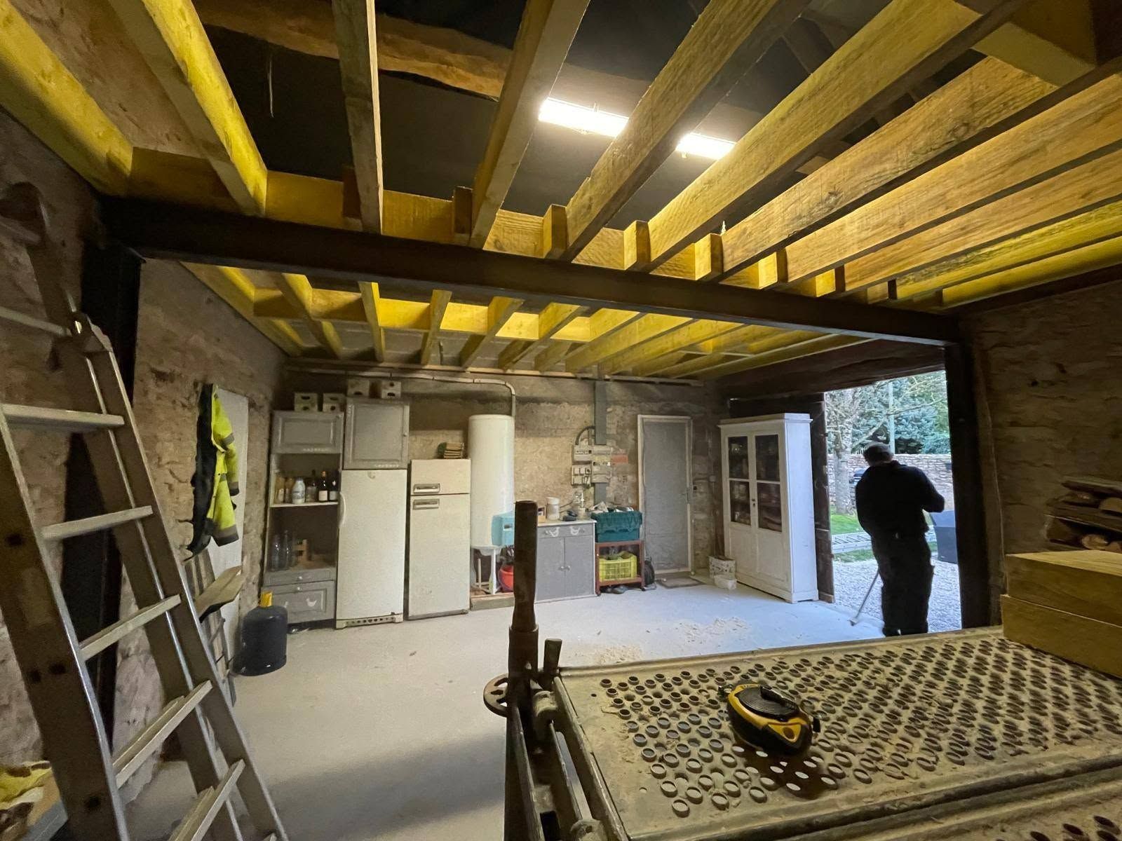 Travaux de rénovation dans une pièce avec des poutres apparentes, des échelles, des outils et une personne debout