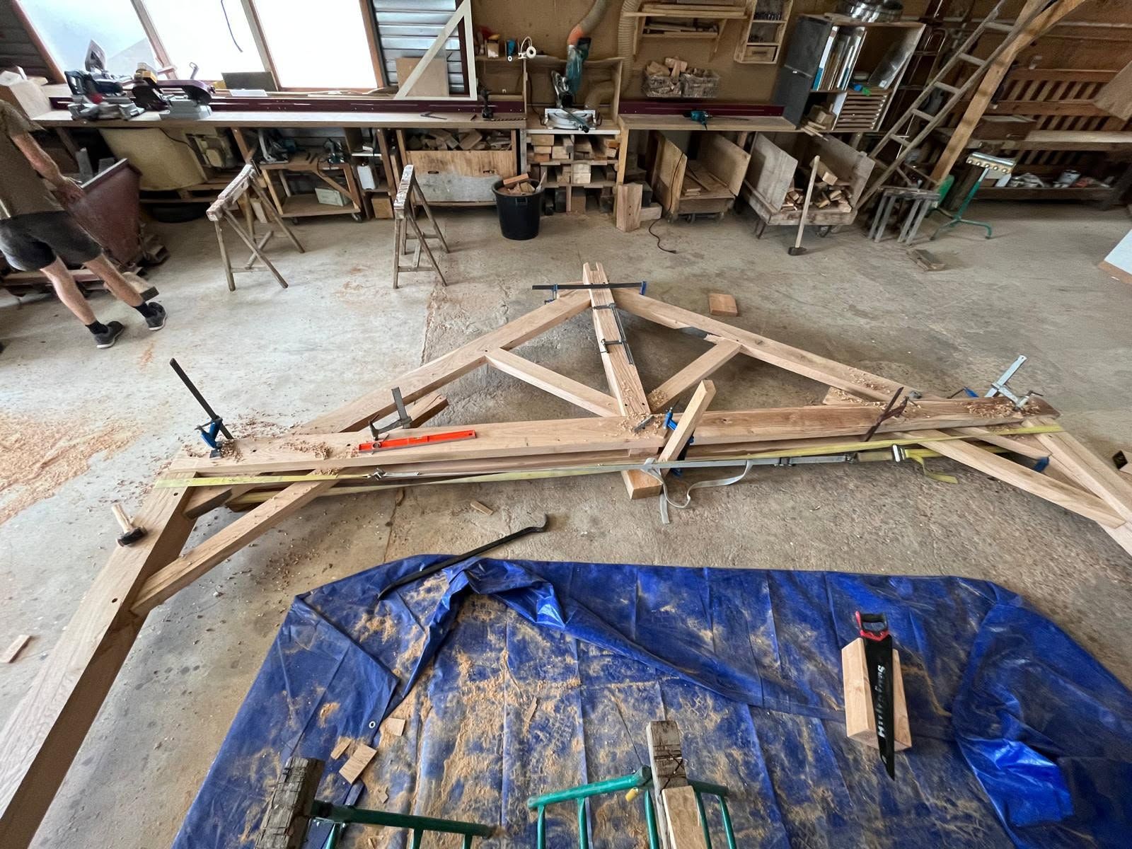 Une ferme de charpente en bois assemblée au sol dans un atelier, avec des outils et une bâche bleue à proximité.