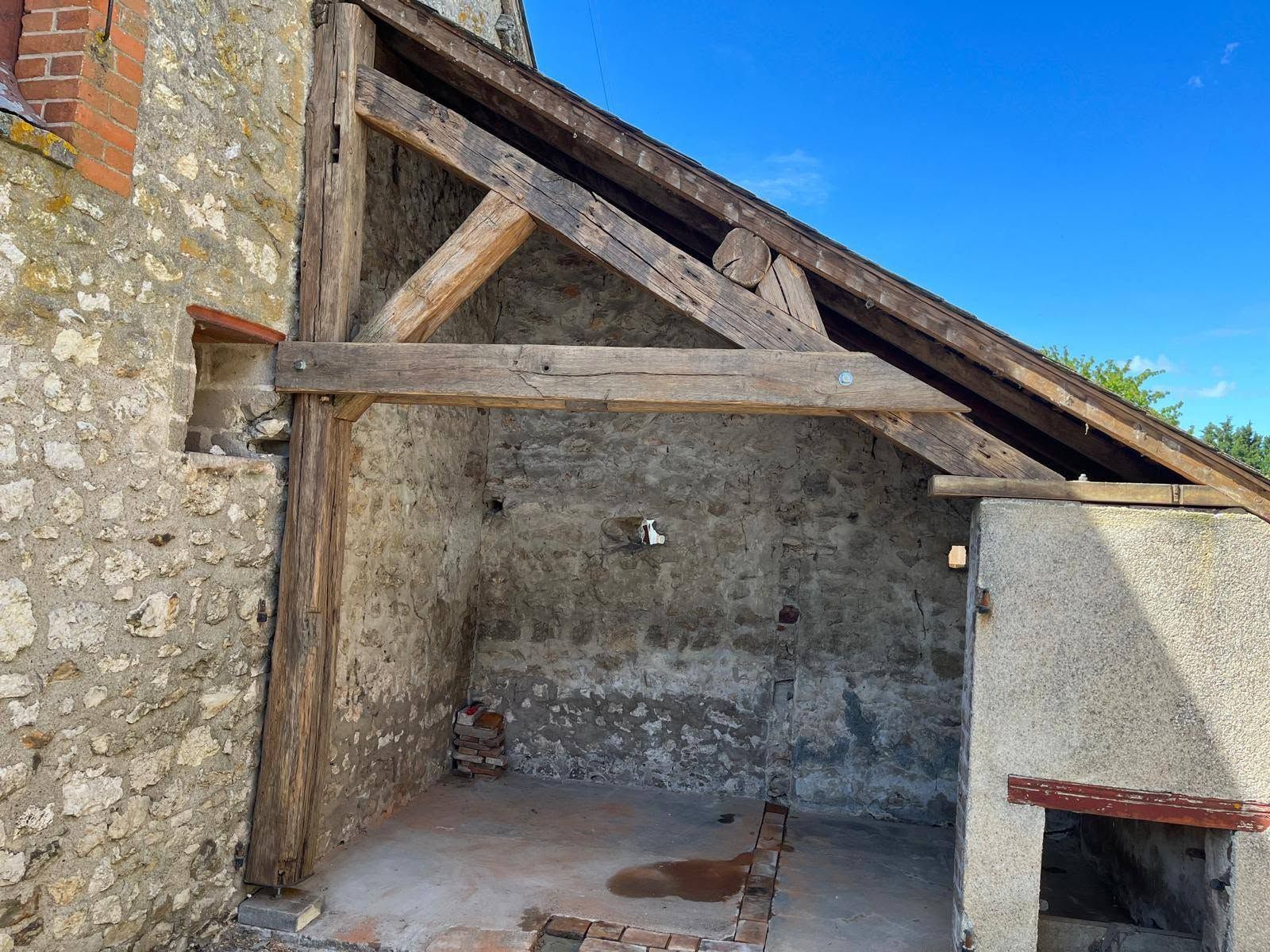 Abri dans une cour en pierre, avec des poutres de toit en bois apparentes et un mur en crépi rugueux sous un ciel bleu