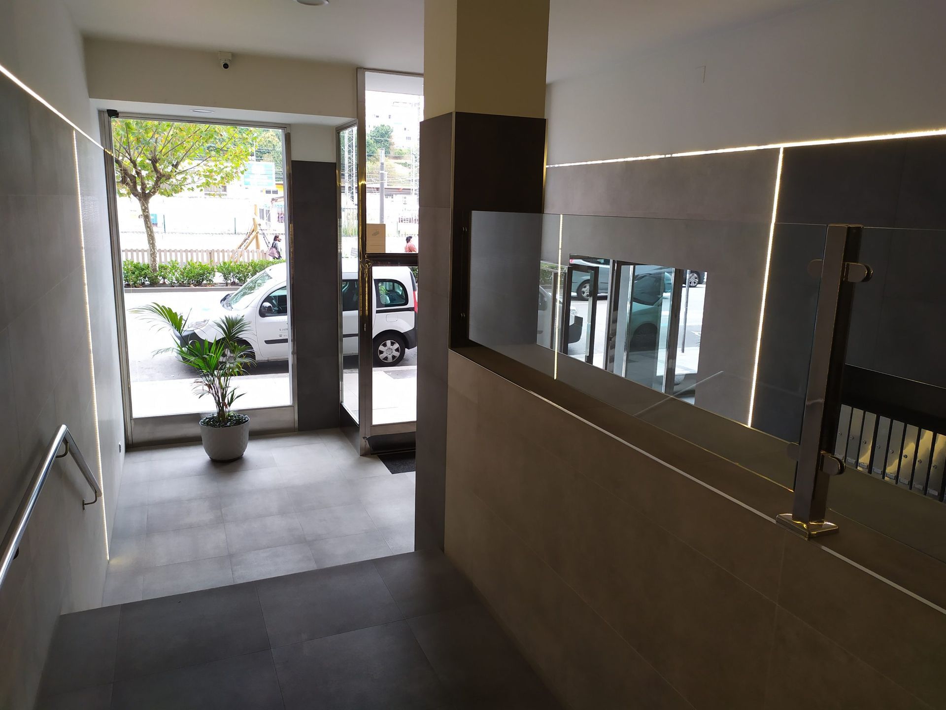 Pasillo con escaleras que conducen a una puerta abierta con vistas al exterior. Azulejos grises, paredes beige y un coche blanco a la vista.