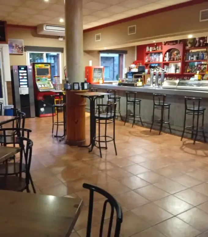Calle estrecha y soleada con mesas y sillas de café frente a pequeños edificios, vista desde una acera de baldosas.