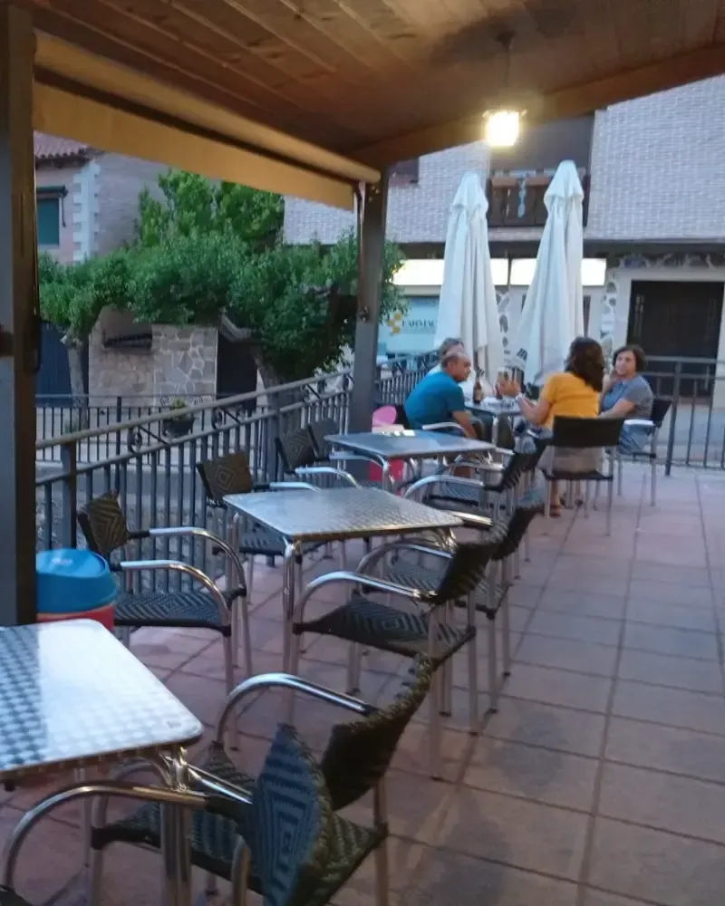 Calle estrecha y soleada con mesas y sillas de café frente a pequeños edificios, vista desde una acera de baldosas.