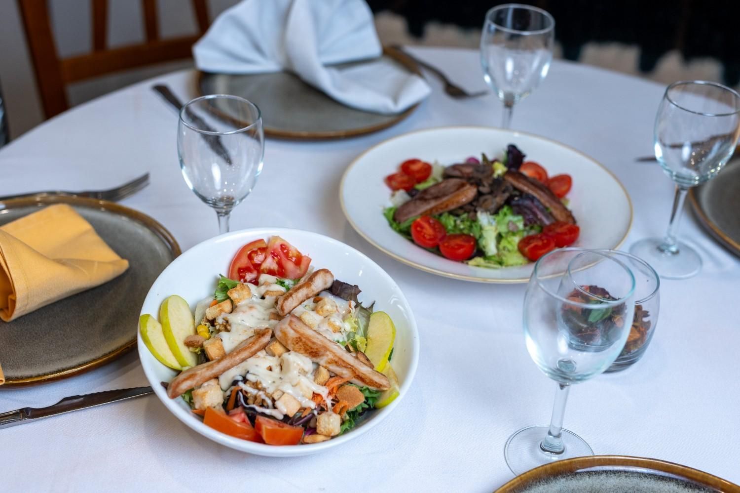 Ensaladas y panecillos servidos en platos sobre una mesa cubierta con un mantel blanco, acompañados de copas de vino.