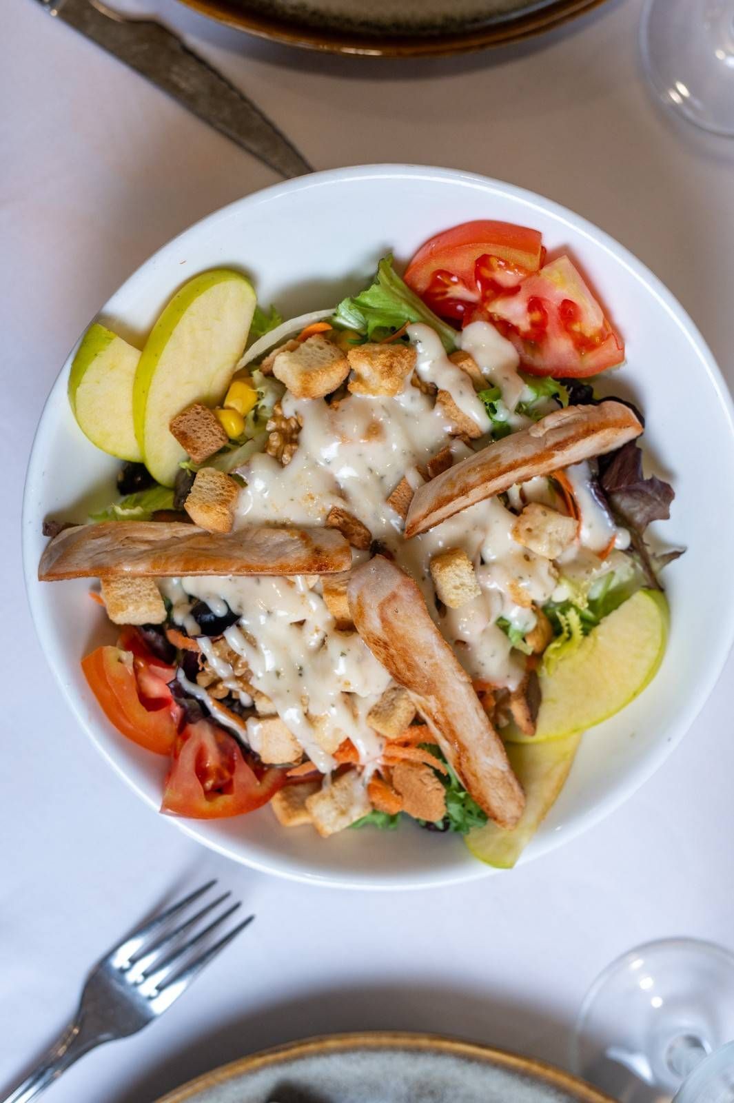 Ensalada de pescado a la parrilla con tomates, rodajas de manzana y queso fundido en un plato blanco.
