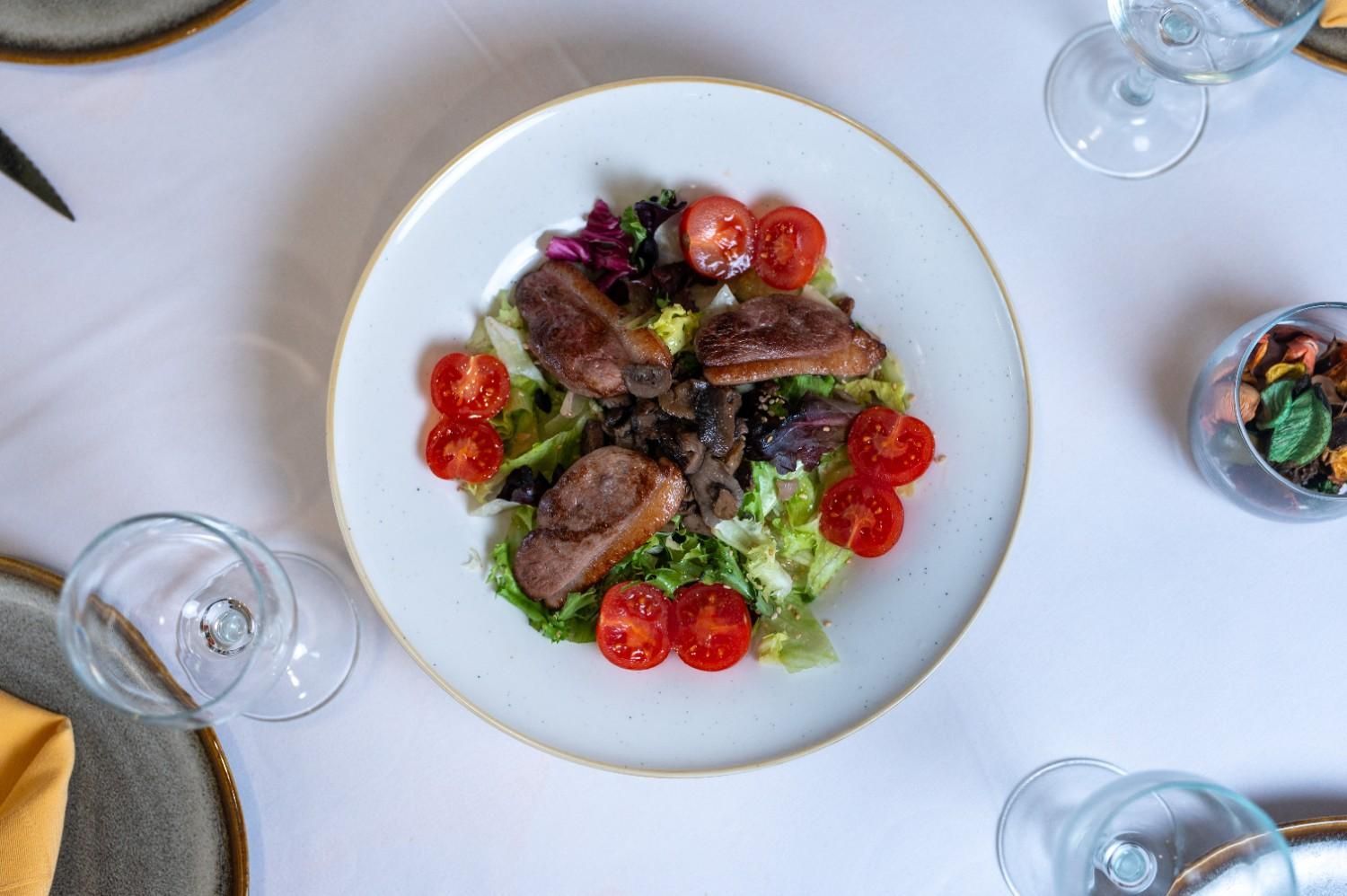 Vista desde arriba de una ensalada de carne con tomates cherry servida en un plato sobre una mesa blanca, rodeada de copas de vino.